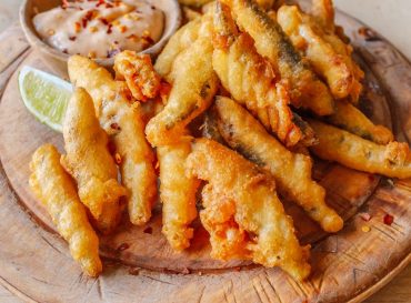Deep fried whitebait sprats served in a crispy golden batter on a wooden platter serving board with a slice of lime and a small dish of chilly sauce as a dip