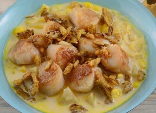 Spicy chowder soup with scallops potatoes and corn and crispy jalapeno pepper topping in blue bowl with golden soup spoon