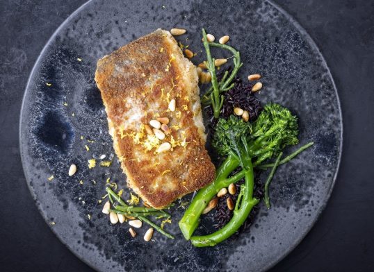 Modern style traditional pan-fried skrei cod fish filet in breadcrumbs with baby broccoli, black rice and roasted pine nuts served as top view on ceramic design plate with copy space
